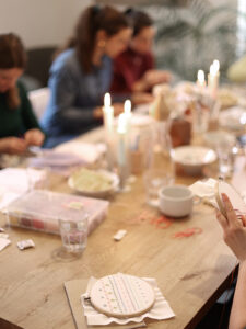 Teilnehmerinnen an einem hölzernen Tisch bei einem Stick-Workshop, beleuchtet von warmem Kerzenschein. Im Vordergrund ein Stickrahmen mit bunten Muster-Stichen. | we love handmade