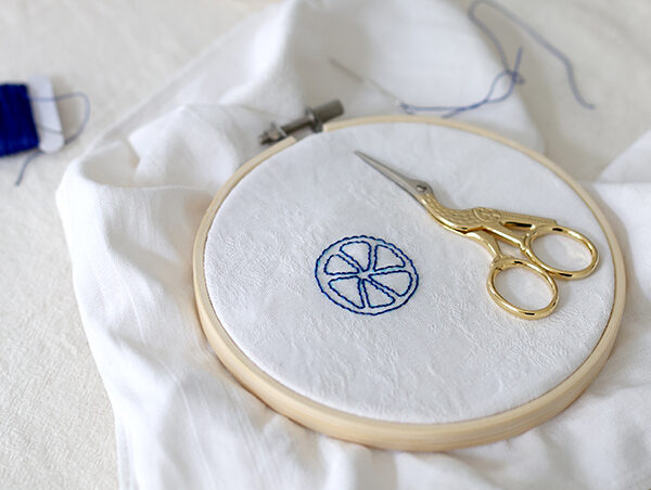 Stickrahmen mit einer blau gestickten Zitronenscheibe auf weißem Stoff, daneben eine goldene Storchenschere und eine blaue Stabkerze – Fokus auf achtsames Handarbeiten und Selfcare. | we love handmade