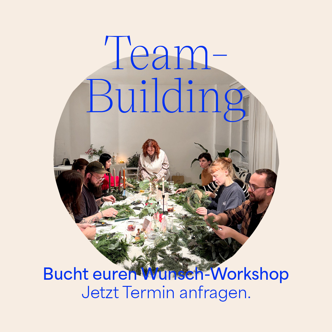 ine gemischte Gruppe von Menschen sitzt gemeinsam an einem großen Tisch in einem hellen Studio und arbeitet konzentriert an grünen Adventskränzen oder Pflanzengestecken. Das Bild ist kreisförmig ausgeschnitten auf beigem Hintergrund. Im Vordergrund steht in markanter blauer Schrift 'Team-Building' sowie der Aufruf 'Bucht euren Wunsch-Workshop. Jetzt Termin anfragen.' | we love handmade