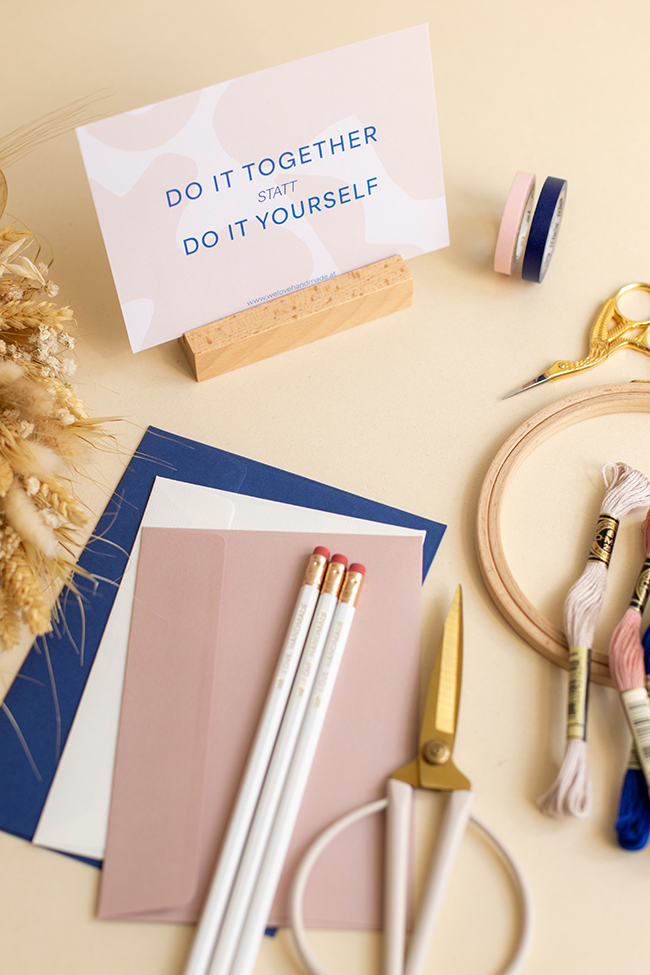 Detailansicht einer Postkarte mit dem Text 'DO IT TOGETHER STATT DO IT YOURSELF' auf einem Holzständer, umgeben von Frühlings-Bastelmaterial.