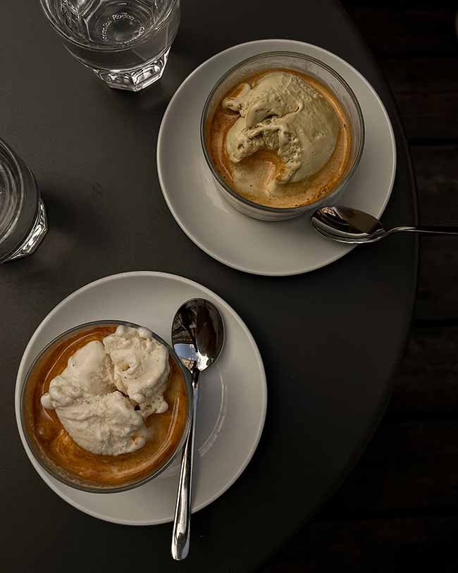 Draufsicht auf zwei Gläser Affogato mit Vanilleeis und Espresso auf einem dunklen Bistrotisch.