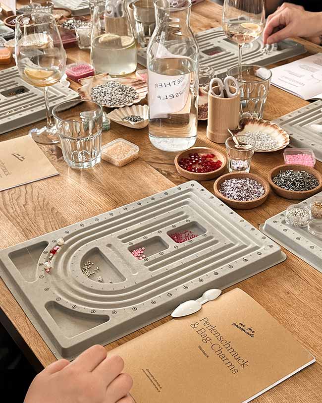 Holz-Workshoptisch mit Perlen-Sortierbrettern, Gläsern mit kühlen Drinks, Muscheln voller Perlen und dem Perlenschmuck-Workbook.