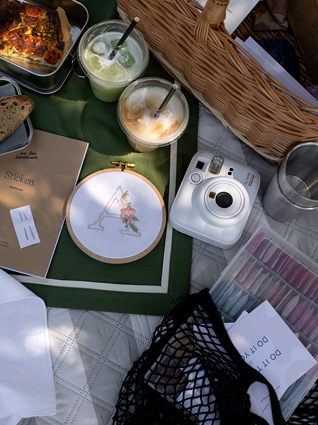 Stickrahmen mit besticktem Monogramm A und Blüten, daneben Instax Kamera, sortierte Stickgarne und eine Netztasche mit Do it together Aufdruck