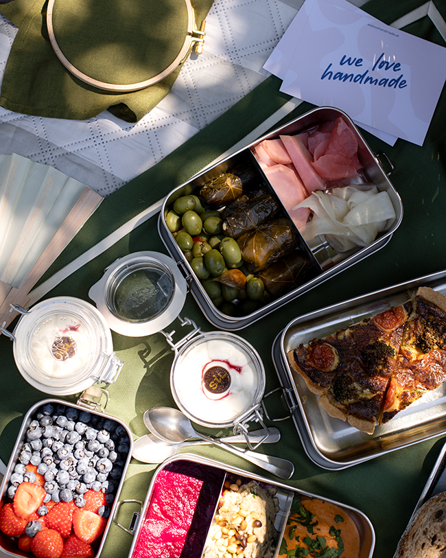 Edelstahldosen mit Oliven, gefüllten Weinblättern, eingelegtem Gemüse, Quiche, Erdbeeren, Heidelbeeren und Trifle im Glas auf grünem Tischtuch