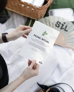 Hände halten die Picnic Ritual Karte von Café SiL vor einem Weidenkorb und einem SiL Fächer auf einer weißen Picknickdecke