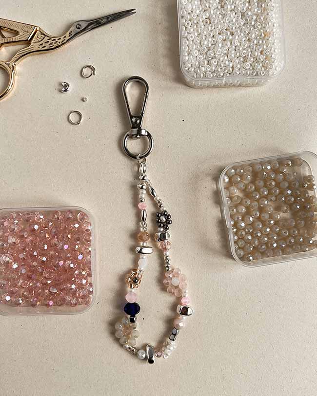 Handgemachter Bag Charm aus bunten Perlen und ein Karabinerhaken, flach auf einem Tisch arrangiert neben kleinen Perlenbehältern und einer Bastelschere.