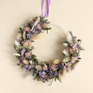 Ein moderner Flowerhoop auf einem weißen Metallring, gebunden mit Trockenblumen in Rosa, Lila und Naturtönen, hängend an einem lila Satinband vor neutralem Hintergrund.