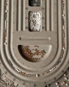 Nahaufnahme von oben auf ein graues Beflockungs-Sortierbrett für Perlenschmuck mit akkurat ausgelegten kleinen Glasperlen und Süßwasserperlen.