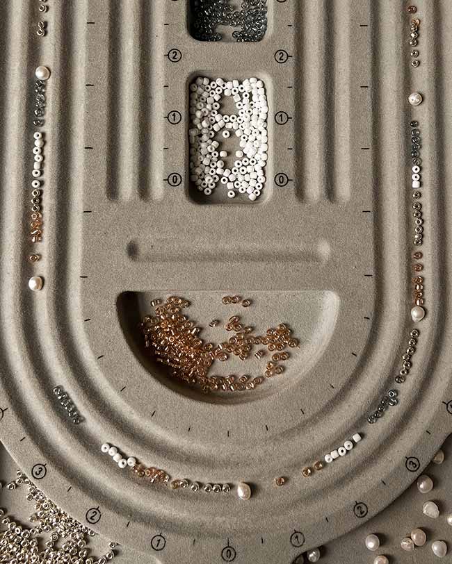 Nahaufnahme von oben auf ein graues Beflockungs-Sortierbrett für Perlenschmuck mit akkurat ausgelegten kleinen Glasperlen und Süßwasserperlen.
