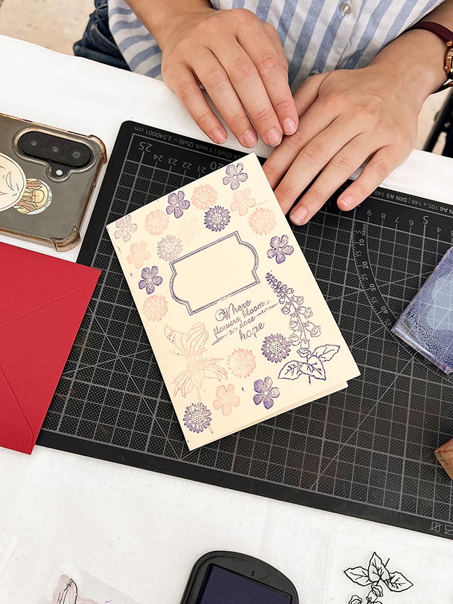 Eine Nahaufnahme einer cremefarbenen Karte, bestempelt mit verschiedenen lila und rosa Blumen und dem Textstempel "Where flowers bloom, so does hope". Hände halten die Karte auf einer Schneidematte.