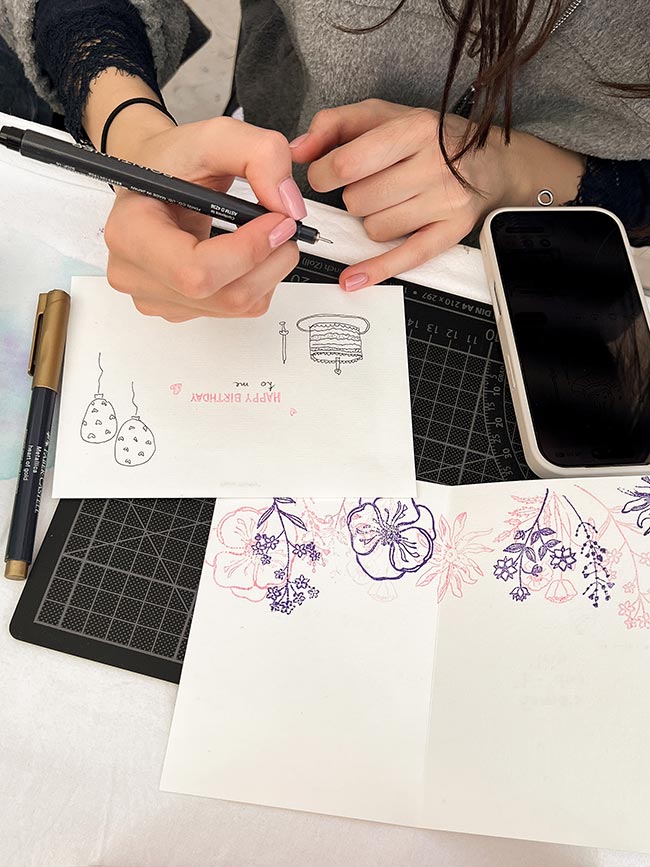 Eine Nahaufnahme einer Hand, die einen schwarzen Fineliner hält, über zwei bestempelten Karten. Eine Karte zeigt Luftballons, die andere bunte Blütenmotive. Im Hintergrund ein Smartphone.