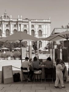 Schwarzweiß-Foto des we love handmade Craft Club Standes beim WAMP Designmarkt vor dem MuseumsQuartier Wien. Drei Personen sitzen am Tisch unter einem weißen Sonnenschirm und basteln, ein Holzaufsteller steht im Vordergrund.