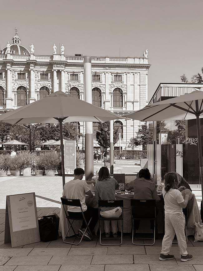 Schwarzweiß-Foto des We Love Handmade Craft Club Standes beim WAMP Designmarkt vor dem MuseumsQuartier Wien. Drei Personen sitzen am Tisch unter einem weißen Sonnenschirm und basteln, ein Holzaufsteller steht im Vordergrund.