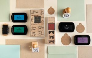 Produktübersicht für DIY-Weihnachtskarten: Holzstempel-Sets (Motive: Weihnachtsgrüße, Schleifen), bunte Stempelkissen, Kraftpapier-Anhänger und farbige Klappkarten mit Umschlägen auf hellem Untergrund.