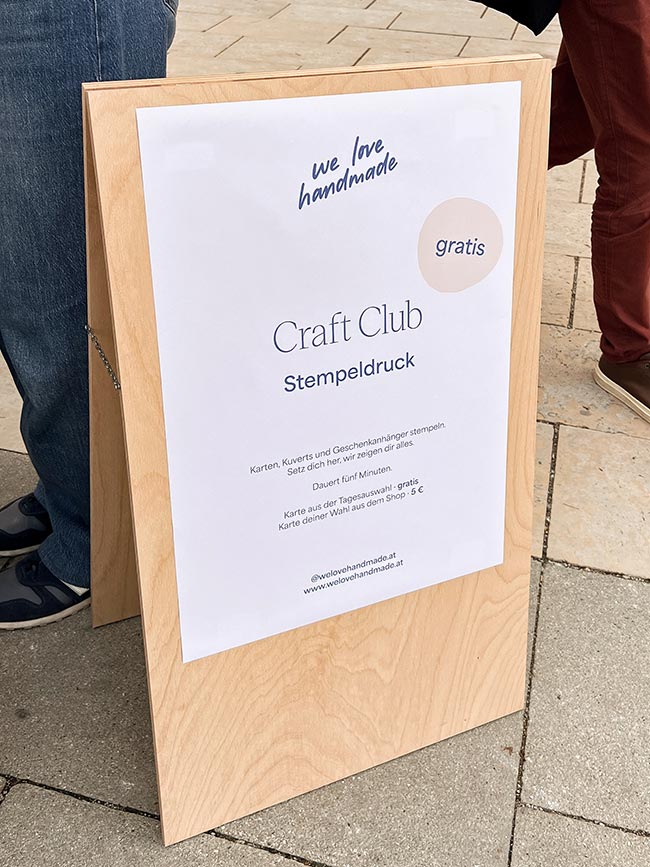 Ein hölzerner Kundenstopper-Aufsteller mit einem Plakat, das für den "Craft Club Stempeldruck" wirbt. Text: "gratis", "Karten, Kuverts... stempeln", "we love handmade". Eine Person steht daneben.