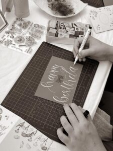 Eine Person schreibt mit einem weißen Faber-Castell Brush Pen "happy birthday" in Kalligrafie auf eine graue Karte, die auf einer Schneidematte liegt, umgeben von Stempeln. (Bild in Schwarzweiß)
