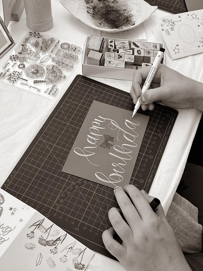 Eine Person schreibt mit einem weißen Faber-Castell Brush Pen "happy birthday" in Kalligrafie auf eine graue Karte, die auf einer Schneidematte liegt, umgeben von Stempeln. (Bild in Schwarzweiß)