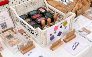Nahaufnahme eines weißen Klappkastens voll mit Stempelkissen und Stempeln auf dem we love handmade Stand. Dahinter Holzboxen mit Farben und Flyer für DIY-Workshops.