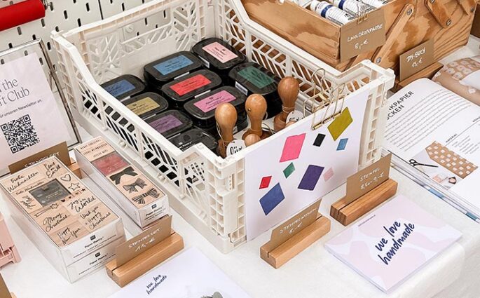 Nahaufnahme eines weißen Klappkastens voll mit Stempelkissen und Stempeln auf dem we love handmade Stand. Dahinter Holzboxen mit Farben und Flyer für DIY-Workshops.