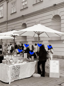 Schwarzweiß-Übersichtsfoto des we love handmade Standes beim WAMP Designmarkt. Mehrere Personen stehen am Verkaufstisch, eine lange Schlange ist im Hintergrund zu sehen. Weiße Sonnenschirme und die MQ-Fassade.