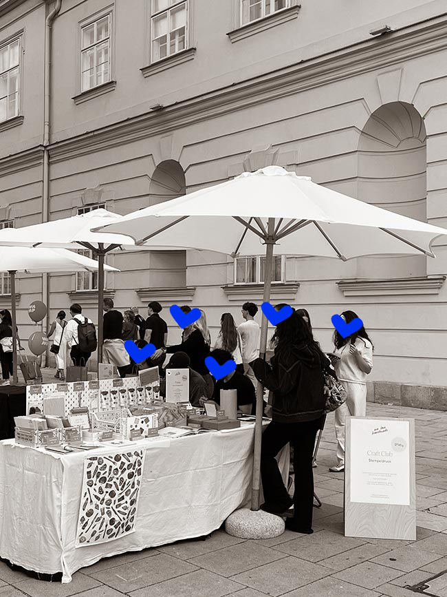 Schwarzweiß-Übersichtsfoto des We Love Handmade Standes beim WAMP Designmarkt. Mehrere Personen stehen am Verkaufstisch, eine lange Schlange ist im Hintergrund zu sehen. Weiße Sonnenschirme und die MQ-Fassade.
