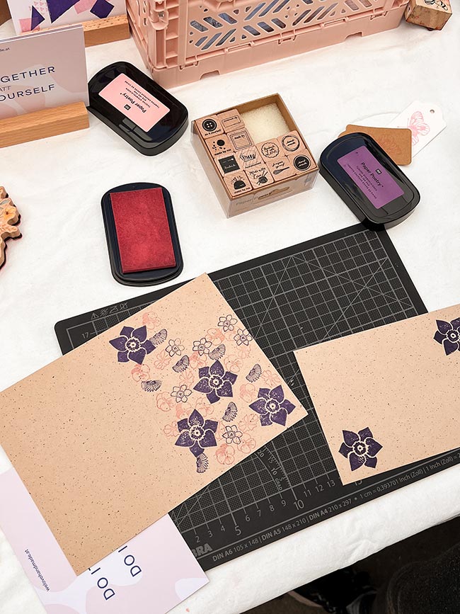 Nahaufnahme von Stempelkissen, Holzstempeln und einer mit lila Blütenmotiven bestempelten Karte auf einer Schneidematte beim We Love Handmade Craft Club Stand.