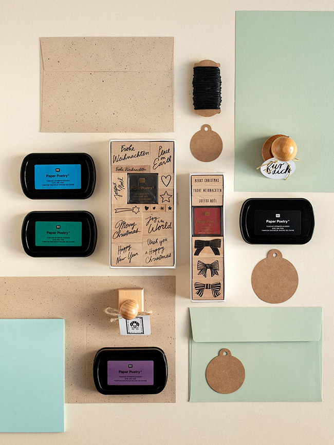 Kreativ-Set für Weihnachtspost: Geöffnete Holzstempel-Kästen, Stempelkissen (Blau, Grün, Schwarz, Lila), Anhänger aus Kraftpapier und hellgrüne Umschläge, bereit zum Stempeln.