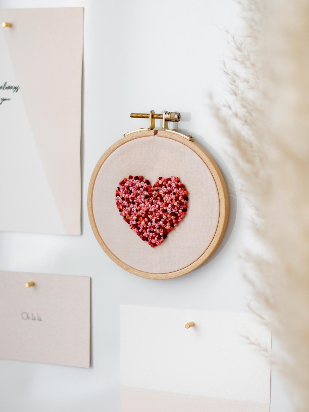 Gesticktes Herz aus roten und rosa Knötchenstichen in einem runden Holzstickrahmen an einer hellen Wand
