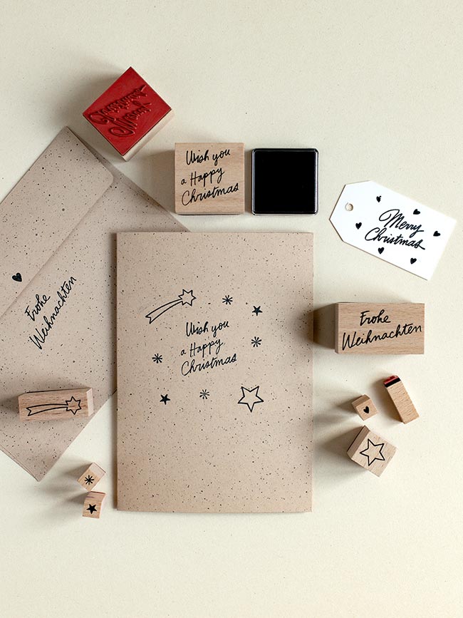 Gestempelte Klappkarte aus Recycling-Kraftpapier mit Sternen, Sternschnuppe und Schriftzug „Wish you a Happy Christmas“, umgeben von Holzstempeln und Stempelkissen.
