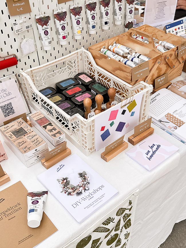 Nahaufnahme eines weißen Klappkastens voll mit Stempelkissen und Stempeln auf dem We Love Handmade Stand. Dahinter Holzboxen mit Farben und Flyer für DIY-Workshops.