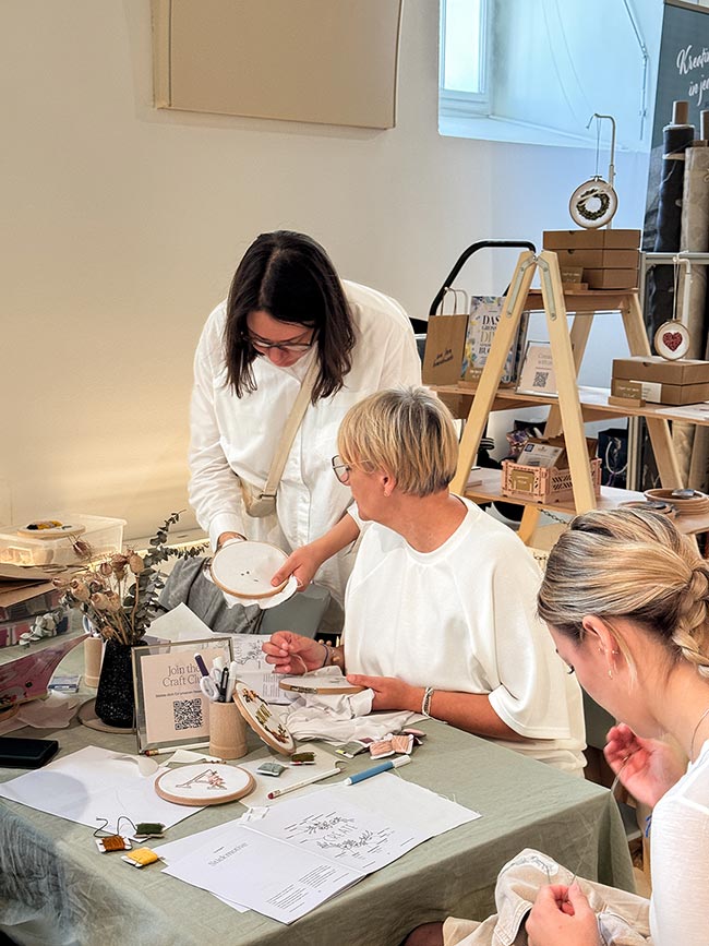 Workshop-Leiterin zeigt einer Teilnehmerin einen Stickrahmen am Craft Club Tisch, dahinter ein Regal mit Stickprodukten