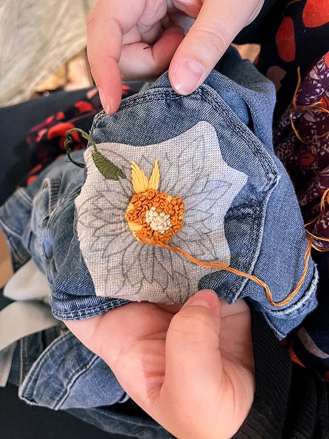 Hände halten eine Jeansjacke mit halb gestickter Sonnenblume in Orange und Gelb auf aufgebügeltem Stickvlies