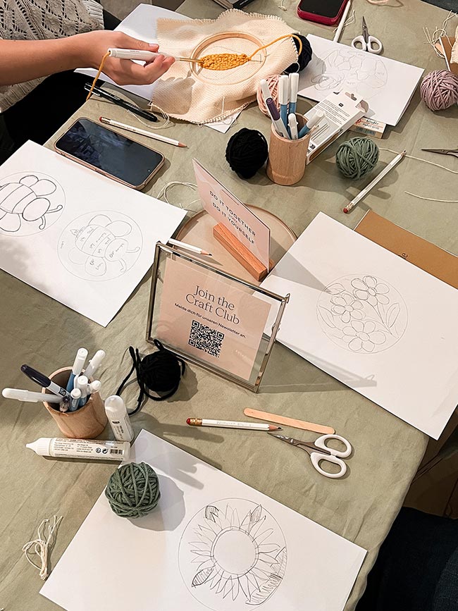 Workshop-Tisch mit Motiv-Skizzen von Sonnenblume und Blüten, Garnknäueln und einem Punch-Needle-Rahmen in Arbeit