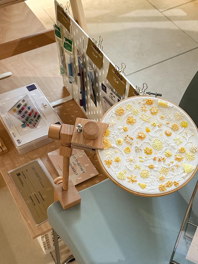 Stickrahmen mit gelbem Blütenmotiv auf einem Holz-Tischständer, dahinter Lochwand mit Sticknadeln und eine Box mit Stickgarnen